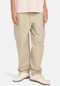 Beige cargobroek met een ontspannen pasvorm, voorzien van zijzakken en een gestructureerde stof. Gecombineerd met witte sneakers en een licht shirt.