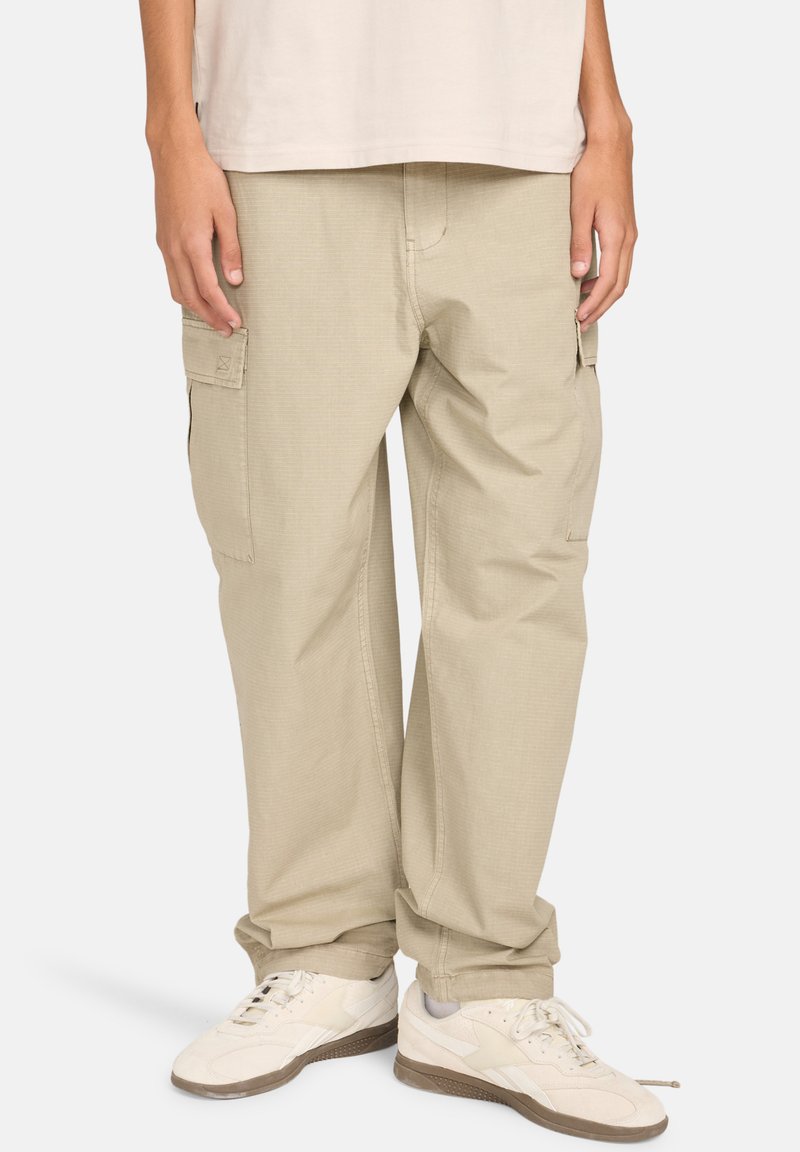Beige cargobroek met een ontspannen pasvorm, voorzien van zijzakken en een gestructureerde stof. Gecombineerd met witte sneakers en een licht shirt.