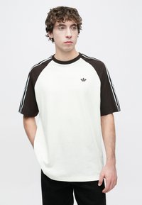 Λευκό και καφέ κοντομάνικο μπλουζάκι με ραγκλάν μανίκια, με τρεις μαύρες ρίγες στους ώμους και ένα μικρό λογότυπο της μάρκας στο στήθος.