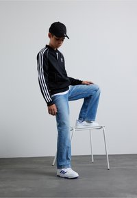 Veste zippée noire avec des rayures blanches, jean bleu en denim et baskets blanches avec des accents bleus. Le mannequin est assis sur un tabouret blanc.