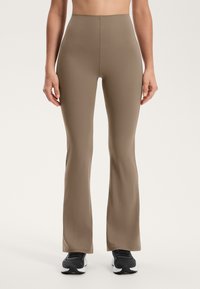 Bež uske flare legginge z visokim pasom, ki imajo gladko teksturo in se tesno prilegajo stegnum, v kombinaciji z črnimi športnimi čevlji.