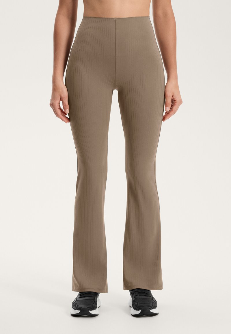 Bež uske flare legginge z visokim pasom, ki imajo gladko teksturo in se tesno prilegajo stegnum, v kombinaciji z črnimi športnimi čevlji.