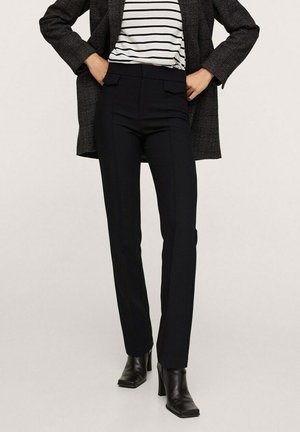 Person trägt schwarze hoch taillierte, schmal geschnittene Hose, schwarze Stiefel mit Absatz, gestreiftes Oberteil und dunklen karierten Blazer mit den Händen in den Taschen.