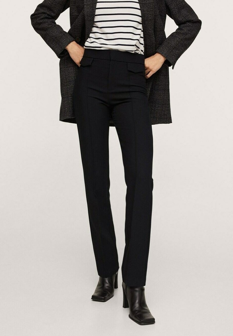 Personne portant un pantalon slim noir taille haute, des bottes noires à talons, un haut rayé et un blazer à carreaux foncé avec les mains dans les poches.