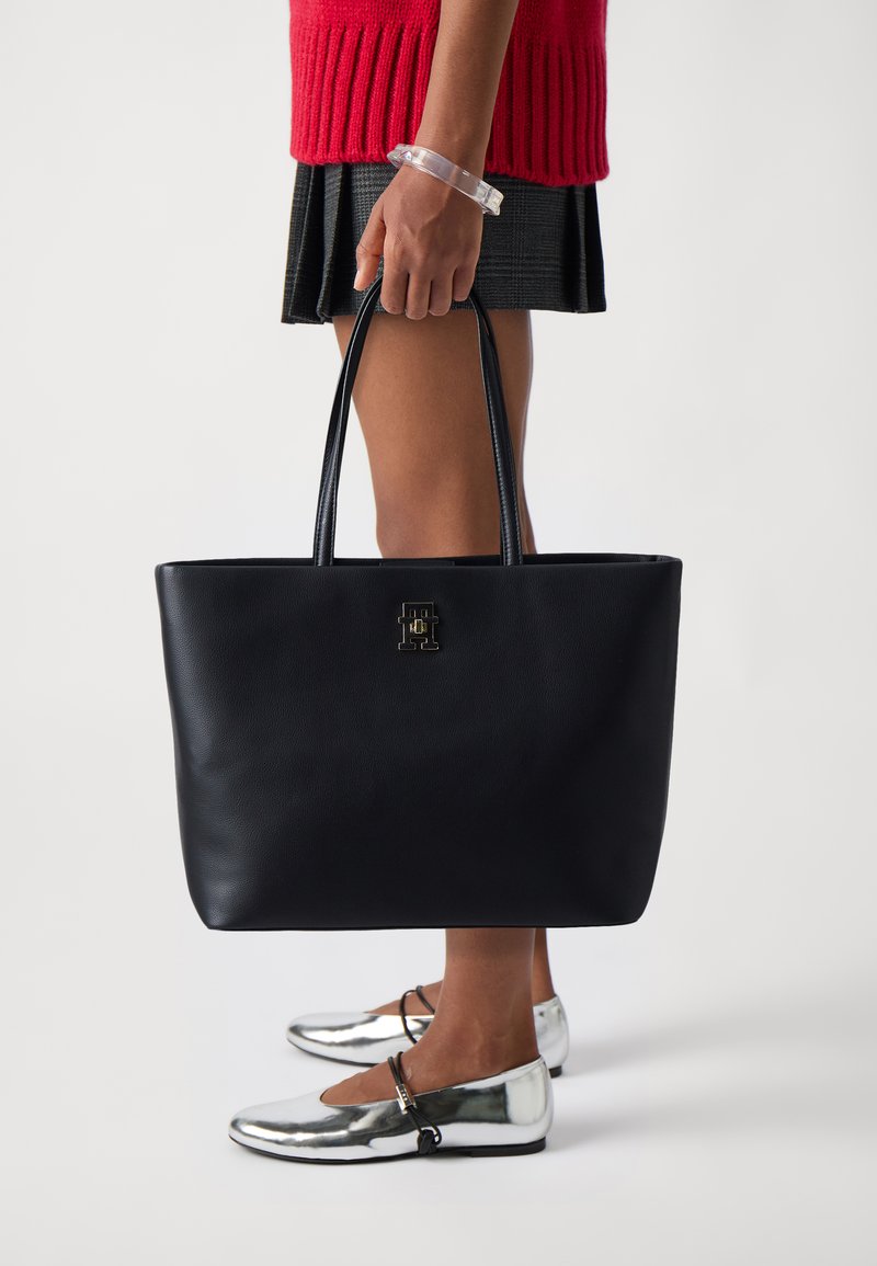 Black leather tote bag with double handles, featuring a gold emblem on the front. Worn with a red sweater and a black skirt. Silver flats.