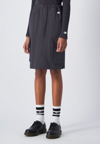 Gonna nera, di lunghezza al ginocchio, con elastico in vita, due tasche laterali e superficie liscia. Indossata con scarpe nere e calze a righe.