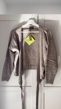 Light brown, cropped cardigan made of soft knit material. Features long sleeves, open front, and ties at the waist. Brand tag visible.