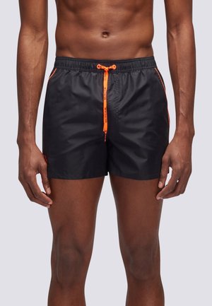 Uomo che indossa pantaloncini da bagno neri con coulisse arancione brillante e bordatura laterale, in piedi contro uno sfondo chiaro e uniforme.