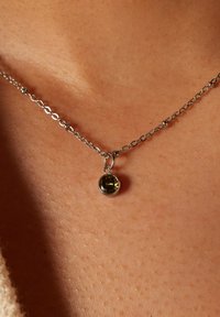 Zilveren ketting met een ronde, getextureerde hanger met een groenachtig gele steen, rustend op de huid. De ketting heeft kleine metalen schakels.