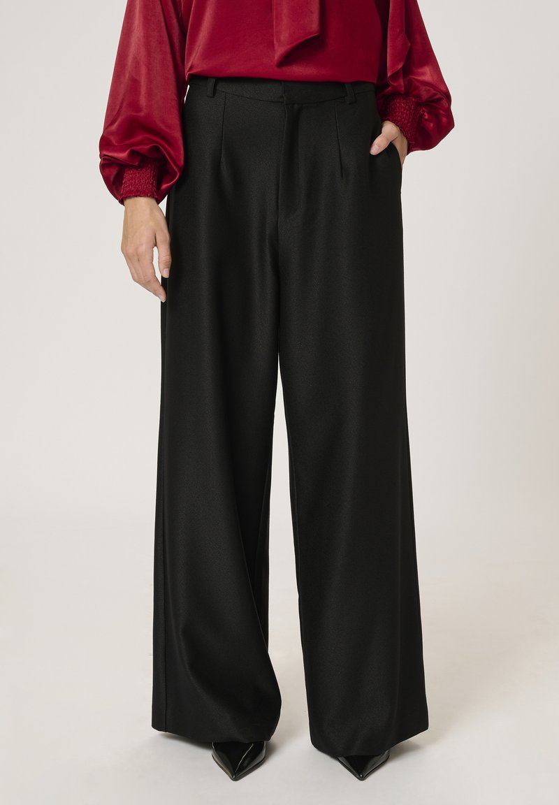 Black wide-leg trousers with a smooth texture, featuring a front crease, tailored fit, and a subtle sheen. Paired with pointed black heels.