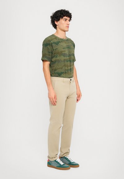 Grön kamouflage t-shirt, beige slim-fit byxor och turkosa sneakers med vita snören. T-shirten har ett texturerat kamouflage-mönster.