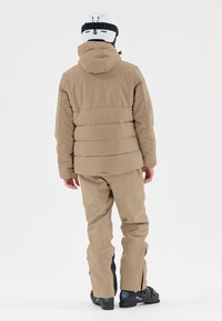 Beige Daunenjacke mit Kapuze, kombiniert mit passenden Hosen. Sie verfügt über ein gepolstertes Design und Reißverschlüsse an den Knöcheln, begleitet von schwarzen Schneestiefeln.