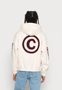 Krémová mikina s kapucí s velkým bordovým kruhovým logem na zádech. Má žebrované manžety a pas, s textem na rukávech. Džíny na spodní část.