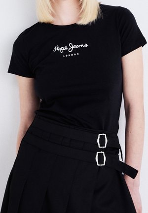 Person iført sort Pepe Jeans London t-shirt og sort højtaljet nederdel med dobbelt sølvspænde, foran ens hvid baggrund.