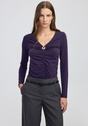 Fioletowa bluzka z długim rękawem z marszczonym detalem z przodu, z okrągłym akcentem metalowym. Noszona z szarymi dopasowanymi spodniami i czarnym paskiem.