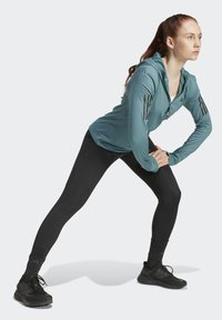 Ljus teal atletisk hoodie med dragkedja framtill och tre svarta ränder på varje ärm, matchad med svarta leggings och svarta sneakers.