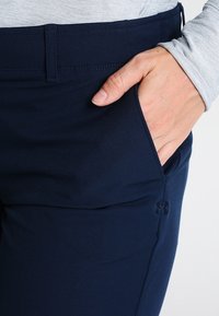 Marinblå byxor i stretchtyg, med en sidficka och en slät yta, samt en synlig söm längs midjebandet.