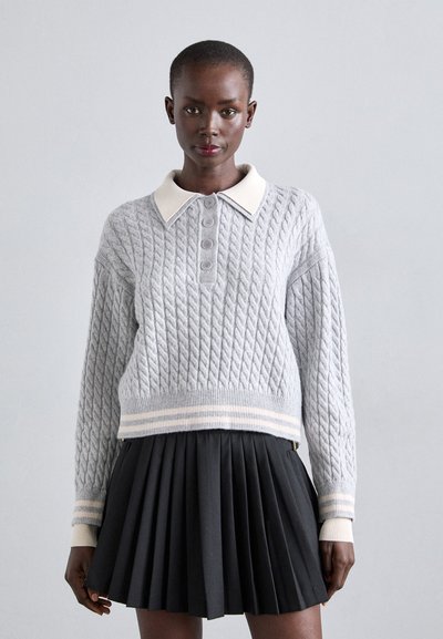 Femme portant un pull en maille torsadée gris clair avec un col et des poignets blancs, associé à une jupe plissée noire, se tenant devant un fond gris.