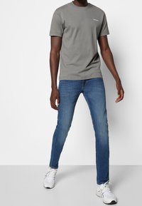 Camiseta de algodón gris con mangas cortas, con un pequeño logo. Combinada con jeans ajustados azules y zapatillas deportivas blancas.