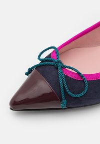 Chaussure plate à bout pointu avec embout bordeaux brillant, corps en daim bleu marine, nœud torsadé vert sarcelle et bordure rose vif autour de l'ouverture.