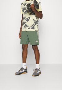 Adidasin graafinen t-paita vaaleassa sävyssä mustilla kuvioilla, vihreät shortsit sivutaskuilla, harmaat urheilukengät oransseilla yksityiskohdilla, pitelee käsipainoa.