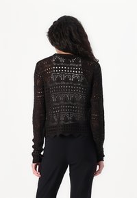 Melnā atvērtā adījumā džemperis ar garām piedurknēm, viļņotu apakšmalu un ģeometriskajiem rakstiem. Aizmugures skats izceļ sarežģītu mežģīņu līdzīgu tekstūru.