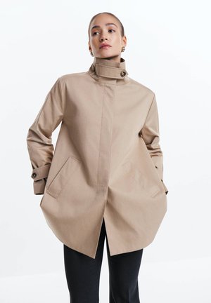 Kvinde iført beige frakke med høj krave, knappede manchetter og skrå lommer, poserende med hænderne i lommerne mod hvid baggrund.