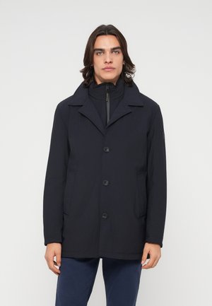 Uomo con capelli scuri fino alle spalle che indossa un cappotto nero abbottonato sopra una giacca con zip e pantaloni blu, in piedi davanti a uno sfondo semplice.