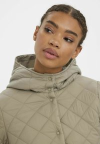 Veste matelassée beige avec capuche, dotée d'une fermeture à boutons-pression et d'une texture en motif diamant. Matériau doux et léger.