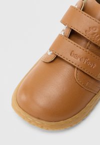 Ταμπά δερμάτινο παπούτσι με δύο λουριά βέλκρο, στρογγυλεμένη μύτη και λαστιχένια σόλα· διαθέτει ανάγλυφο λογότυπο "barefoot" στο πλάι.