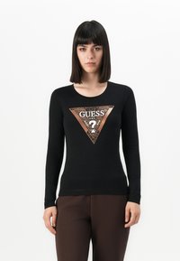 Donna che indossa una maglietta nera a maniche lunghe con un triangolo del logo Guess in stampa leopardata metallica, abbinata a pantaloni marrone scuro, in piedi contro uno sfondo bianco.