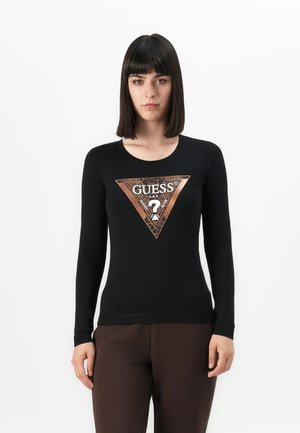 Donna che indossa una maglietta nera a maniche lunghe con un triangolo del logo Guess in stampa leopardata metallica, abbinata a pantaloni marrone scuro, in piedi contro uno sfondo bianco.