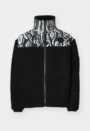 Black fleece jacket with a high collar, featuring a geometric gray and white patterned panel on top and a circular logo detail.