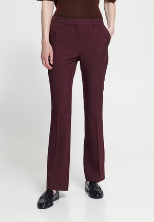 Personne portant un pantalon bordeaux à jambes droites, des mocassins foncés et un haut marron, debout les mains dans les poches devant un fond uni.