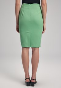 Grön figurnära blyertsskjol med strukturerad tyg, mitt bak slits och en dold dragkedjedetalj i midjan. Svarta klackskor med blockklack synliga.