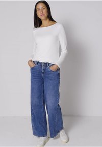 Witte lange mouw top gecombineerd met hooggeprijsde blauwe wijde jeans en witte sneakers. De top heeft een boot-halslijn en aansluitende mouwen.