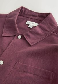 Chemise à manches courtes bordeaux en tissu texturé, avec col plié, fermeture à boutons et poche poitrine. Étiquette visible à l'intérieur.