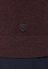 Bordowy sweter z dzianiny z fakturowanym wzorem i czarną skórzaną naszywką z logo na dolnym brzegu. Gładki, ciepły materiał.