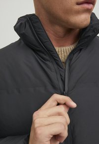 Schwarze Pufferjacke mit hohem Kragen, glatter Textur und einem Reißverschluss vorne. Getragen über einem hellbraunen Strickpullover.