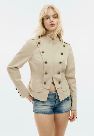 Blond žena nosiaca béžový dvojradový vojenský kabátik s mosadznými gombíkmi a džínsové šortky, stojí s jednou rukou na páse.