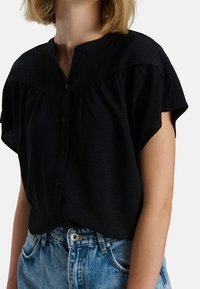 Blusa nera a maniche corte con scollatura rotonda, chiusura a bottoni e dettagli arricciati sulle spalle, abbinata a jeans blu a vita alta.