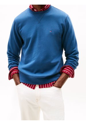 Mężczyzna w niebieskiej bluzie na suwak nałożonej na czerwoną koszulę w paski, biały spodnie, ręce w kieszeniach, na białym tle.