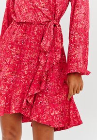 Červené šaty s vzorem, dlouhými rukávy, zavazováním v pase a volánkovým lemem. Materiál má texturovaný povrch s kombinací živých barev.
