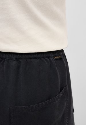 Mustade elastse vöökohaga pükste lähivaade, millel on seljatasku ja väike brändisilt, kantud helevalge üleriidega vöökoha kohal.