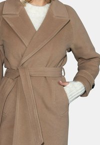 Płaszcz w kolorze camel z dużymi klapami, przewiązany w talii i dwiema bocznymi kieszeniami. Wykonany z miękkiego, teksturowanego materiału, noszony na lekkim swetrze.