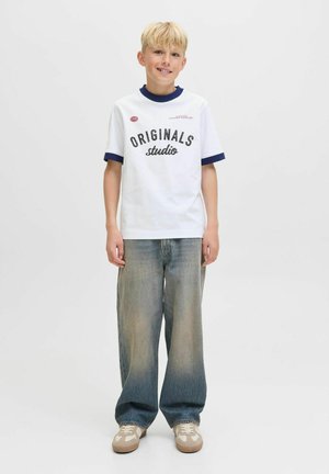 Hvid t-shirt med marineblå ribbet krave og manchetter, med sort tekst "ORIGINALS studio." Sammen med løse blå jeans og beige sneakers.