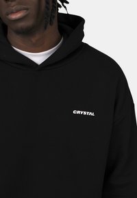 Sweatshirt à capuche noire en tissu doux, arborant un logo "CRYSTAL" blanc sur la poitrine, avec un design simple et décontracté.