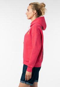 Roze hoodie met een voorste kangoeroezak, witte trekkoorden en geborduurde accenten. Gecombineerd met donkere denim shorts.