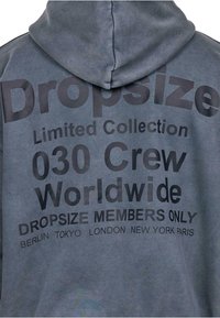 Grau Hoodie mit einer strukturierten Oberfläche. Verziert mit fettem schwarzen Text: "Dropsize Limited Collection 030 Crew Worldwide" und Städtenamen am unteren Rand.