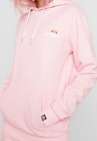Osoba z różowymi włosami nosząca pastelowy różowy hoodie ellesse z kieszenią z przodu i sznurkami, trzymająca ręce w kieszeni, na jednolitym tle.